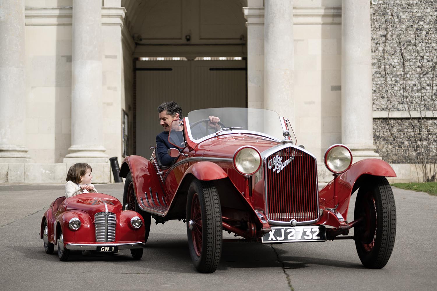 Image of classic red Alfa Romeo car and red J40 pedal car in front of Goodwood House stables